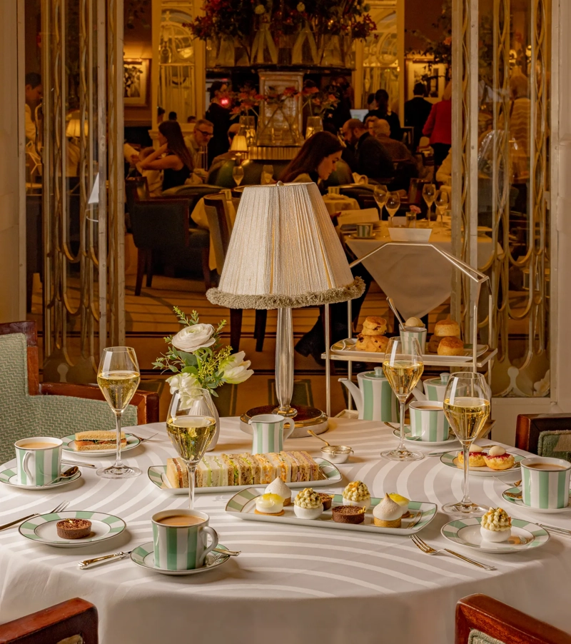 Refined afternoon tea setting with striped china, pastries, and champagne glasses arranged on a round white tablecloth. A softly lit lamp and floral arrangement sit at the center, with guests enjoying tea service in the elegant room beyond.