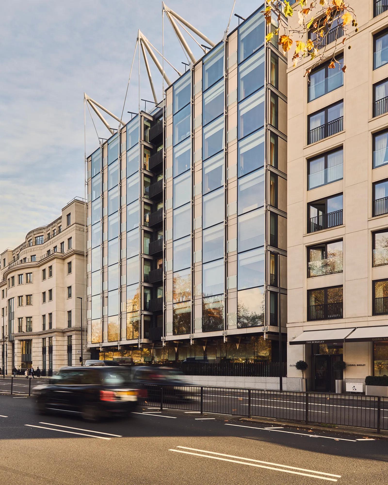 Modern glass-fronted city building beside a busy road, with neighbouring beige buildings and passing traffic.