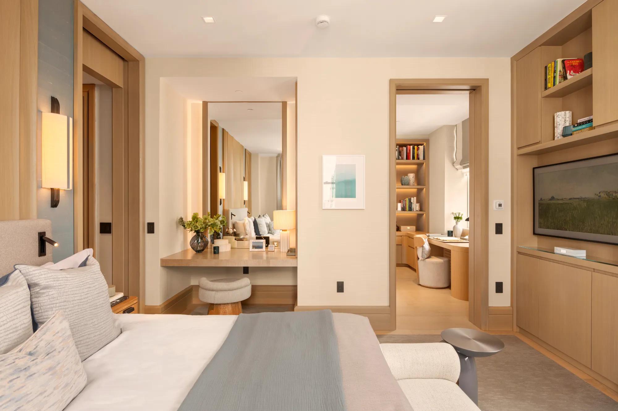 Contemporary bedroom suite with bespoke woodwork, integrated shelving, dressing area and adjoining living space in warm neutral tones.