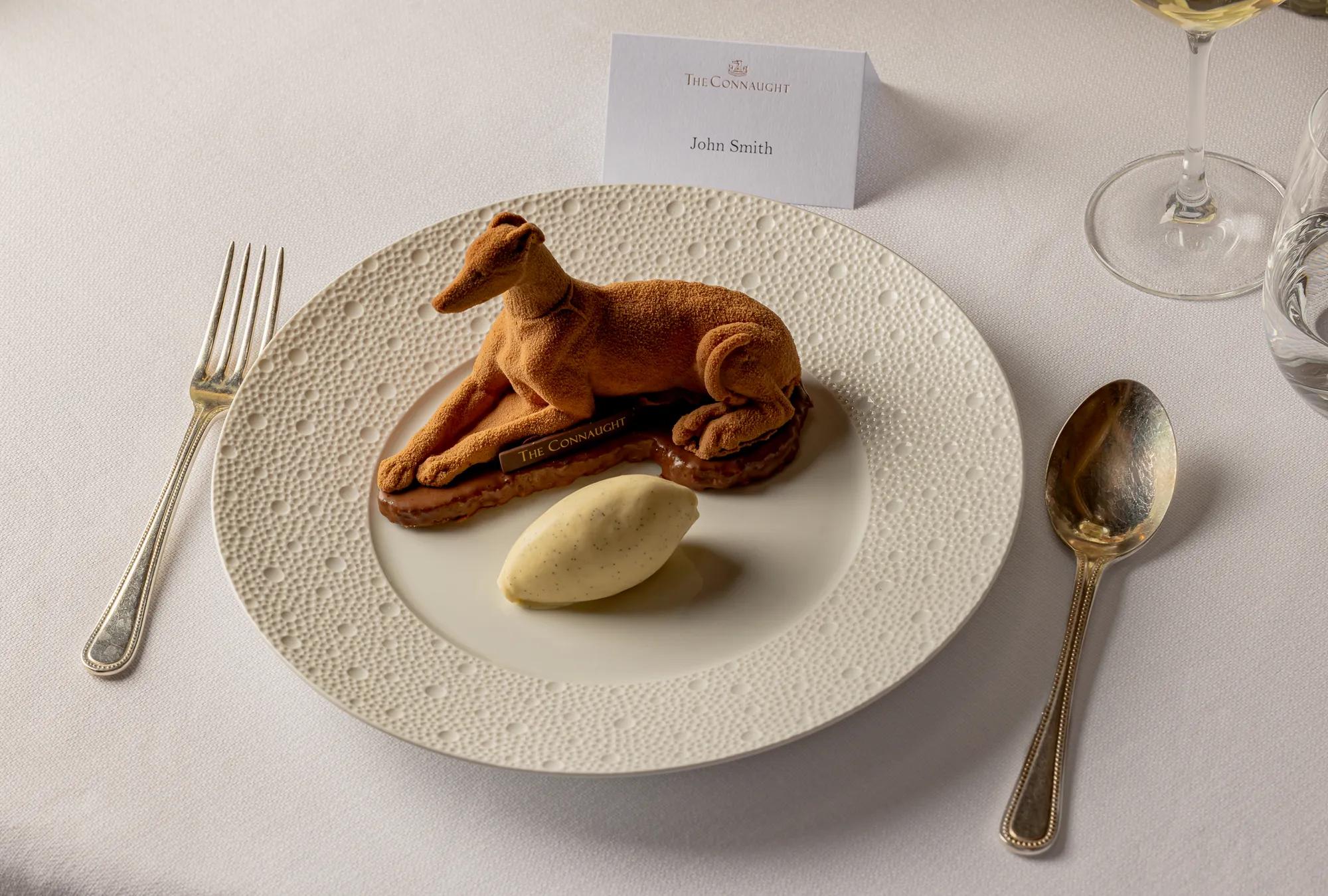 Plated dessert shaped like a reclining dog with a scoop of ice cream on a textured white plate.
