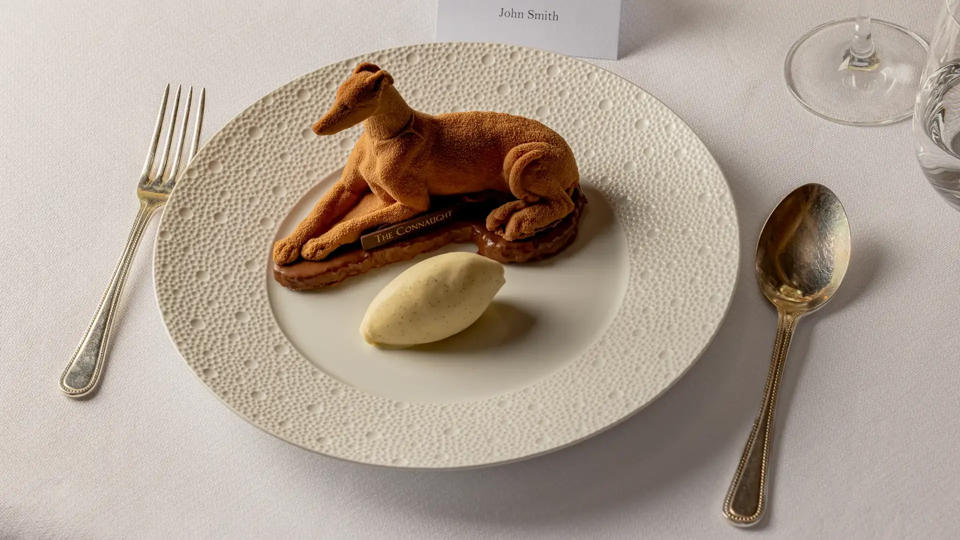 Plated dessert shaped like a reclining dog with a scoop of ice cream on a textured white plate.