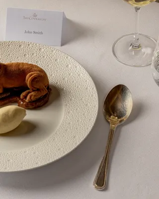 Plated dessert shaped like a reclining dog with a scoop of ice cream on a textured white plate.