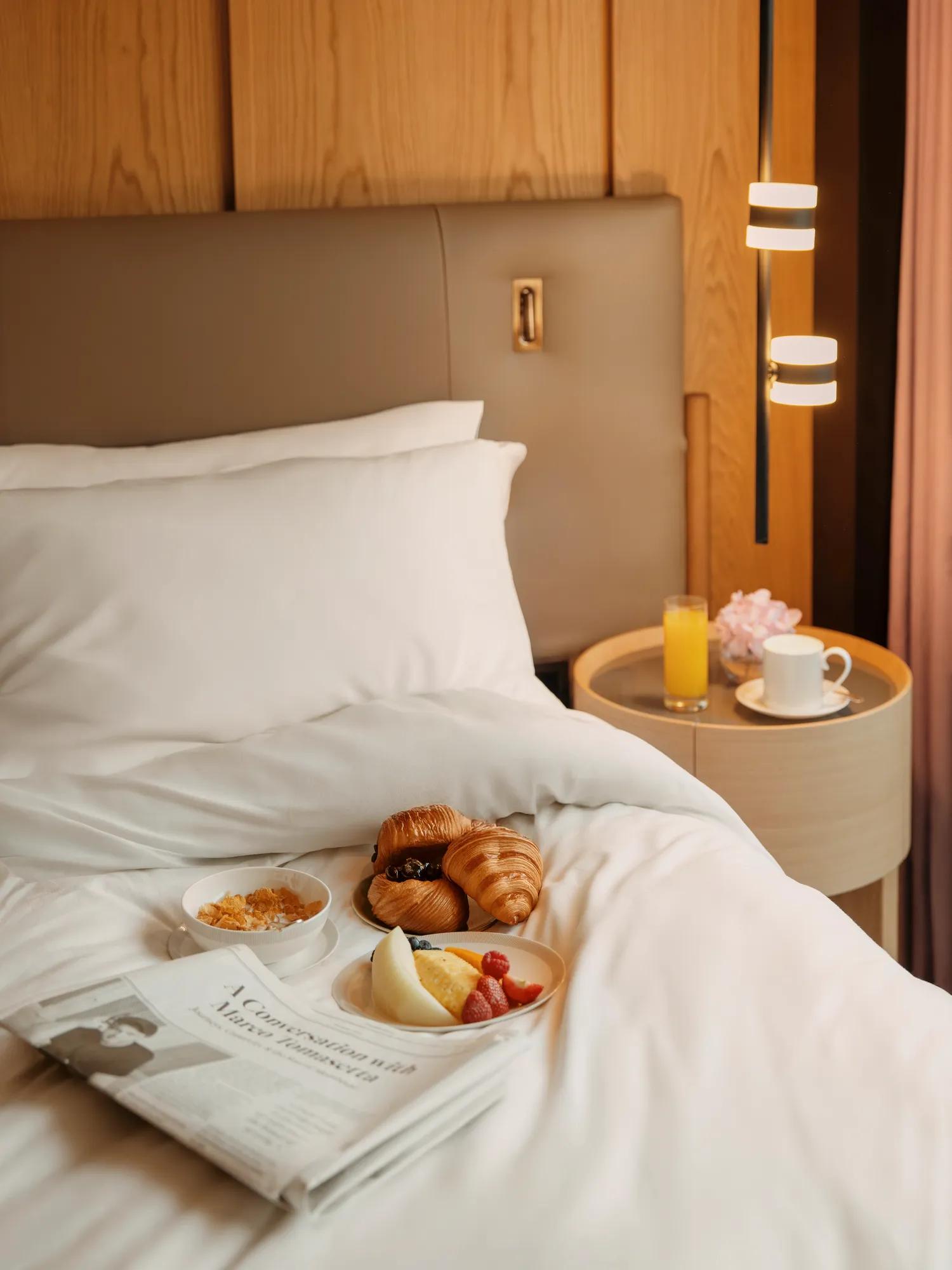 Breakfast tray with croissants, fruit and granola on a neatly made hotel bed, beside a bedside table with coffee, juice and fresh flowers.