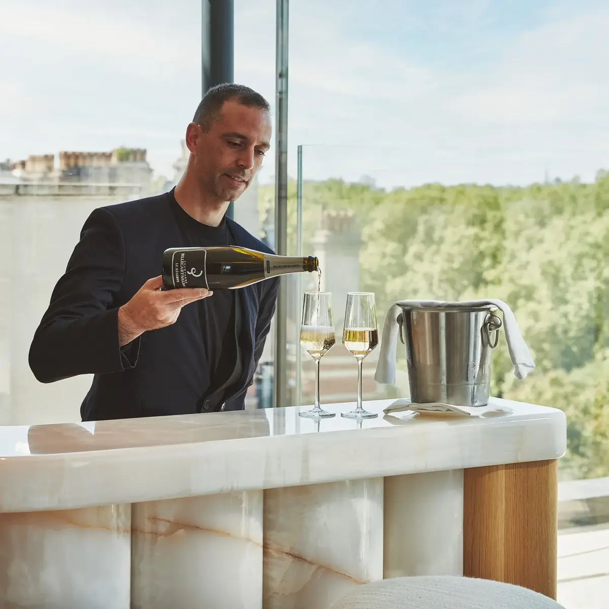 Sommelier pouring white wine into glasses at a bar, with ice bucket and leafy city view beyond
