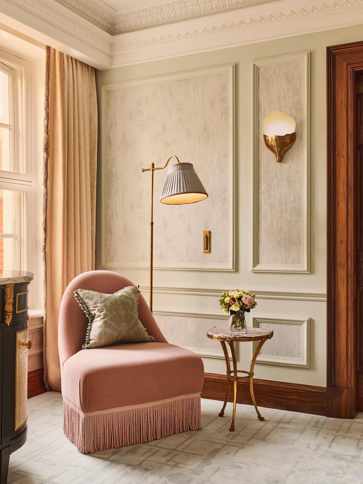 Refined seating corner featuring a blush pink fringed armchair with a patterned cushion, a gold floor lamp, and a small marble-topped side table with fresh flowers beside tall windows.