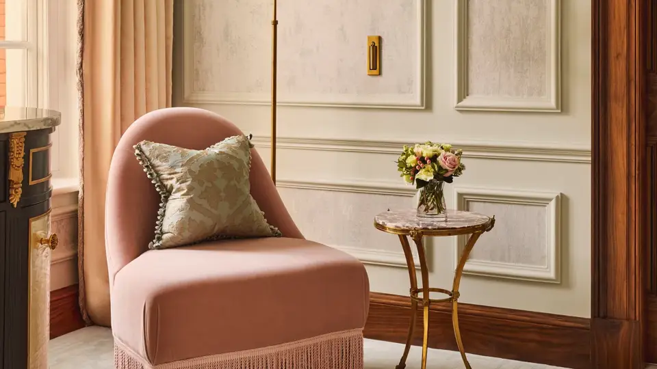 Refined seating corner featuring a blush pink fringed armchair with a patterned cushion, a gold floor lamp, and a small marble-topped side table with fresh flowers beside tall windows.