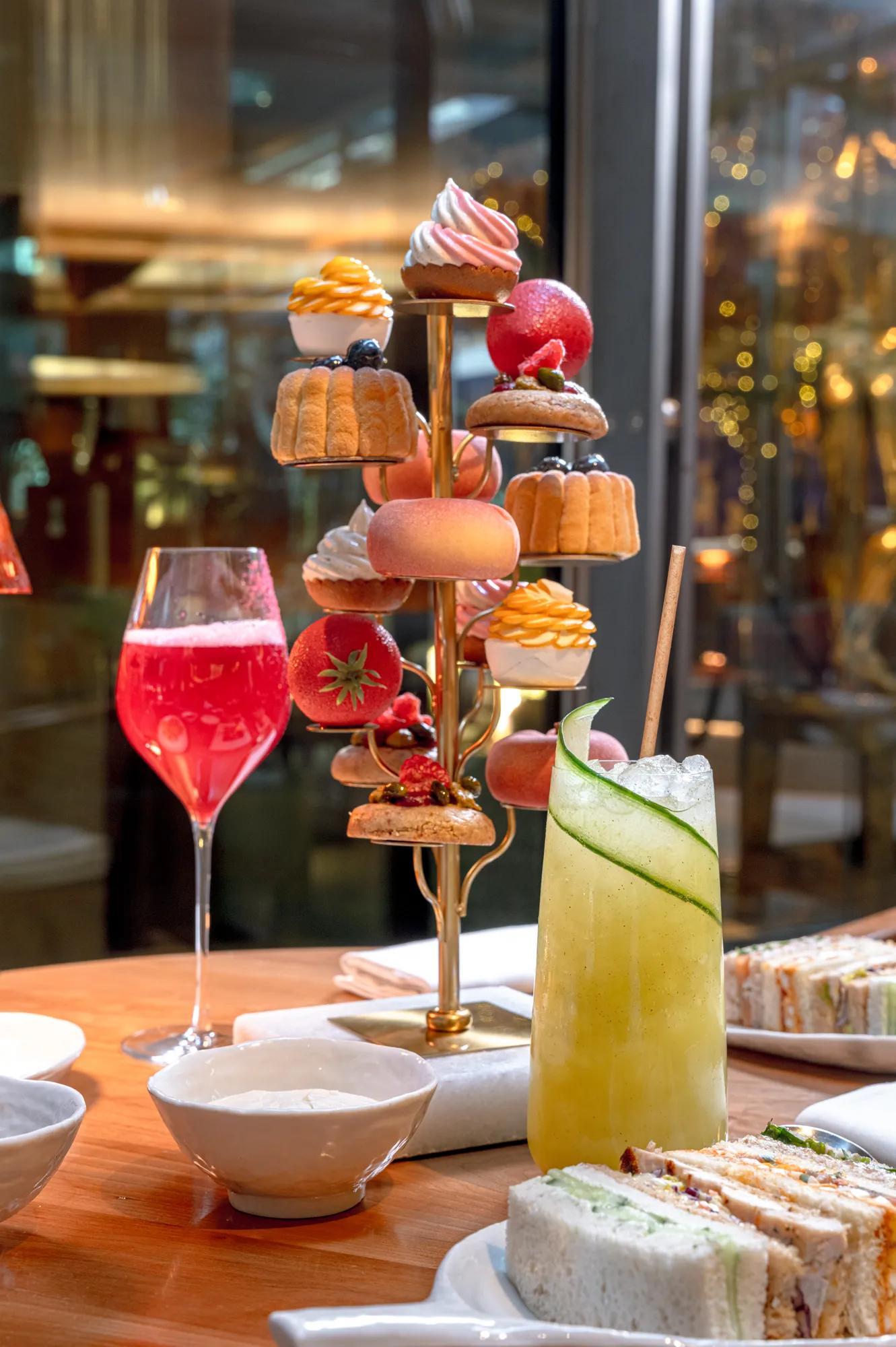 Tiered afternoon tea stand displaying pastries, cakes and fruit desserts beside cocktails and finger sandwiches on a restaurant table with city lights beyond.