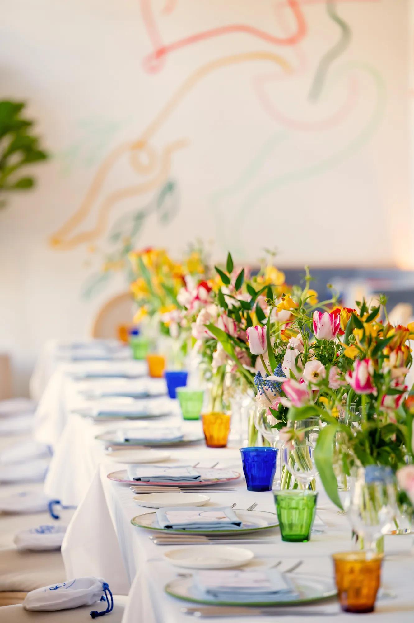 Long private dining table set with colorful glassware and vases of tulips against a mural backdrop.