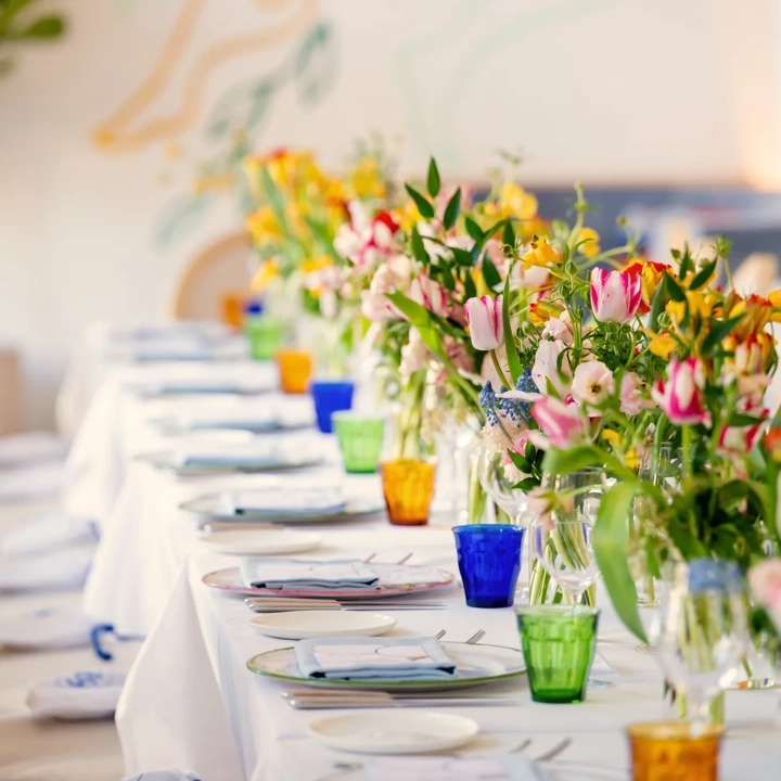 Long private dining table set with colorful glassware and vases of tulips against a mural backdrop.