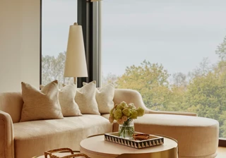 Sunlit living area with curved sofa, brass nesting tables, and large windows overlooking treetops