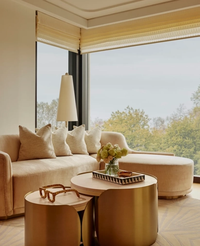 Sunlit living area with curved sofa, brass nesting tables, and large windows overlooking treetops