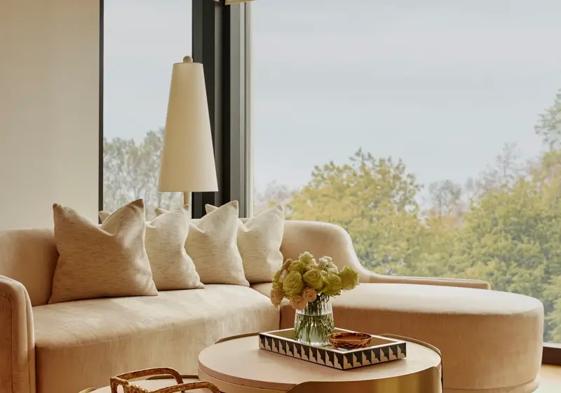Sunlit living area with curved sofa, brass nesting tables, and large windows overlooking treetops