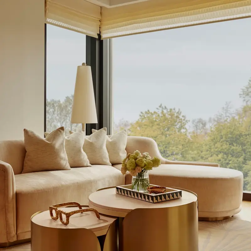 Sunlit living area with curved sofa, brass nesting tables, and large windows overlooking treetops