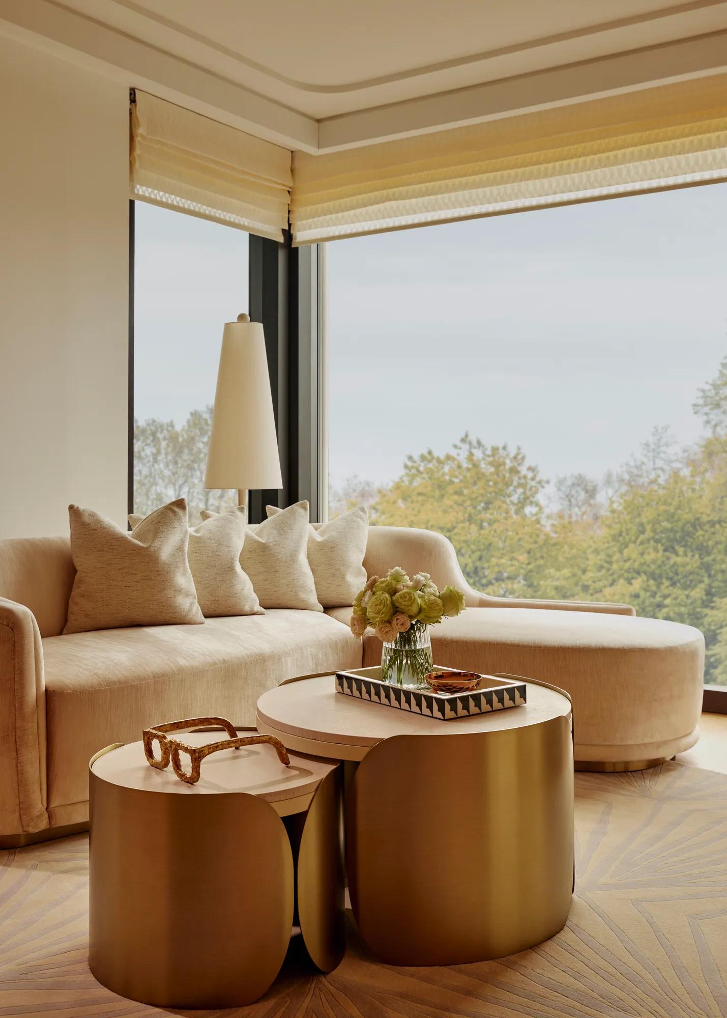 Sunlit living area with curved sofa, brass nesting tables, and large windows overlooking treetops