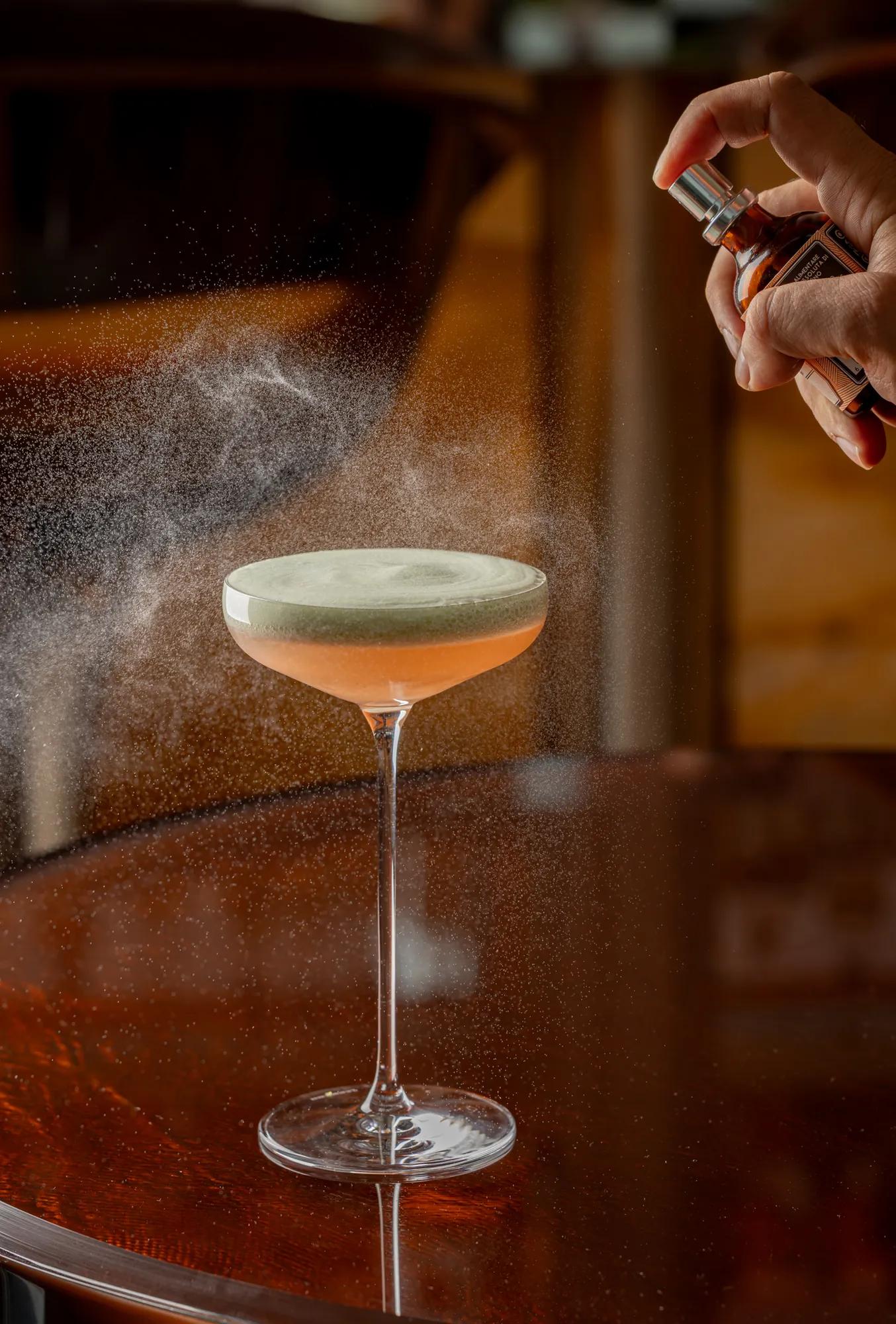 A hand sprays a fine mist over a pale pink cocktail in a coupe glass on a polished wooden bar.