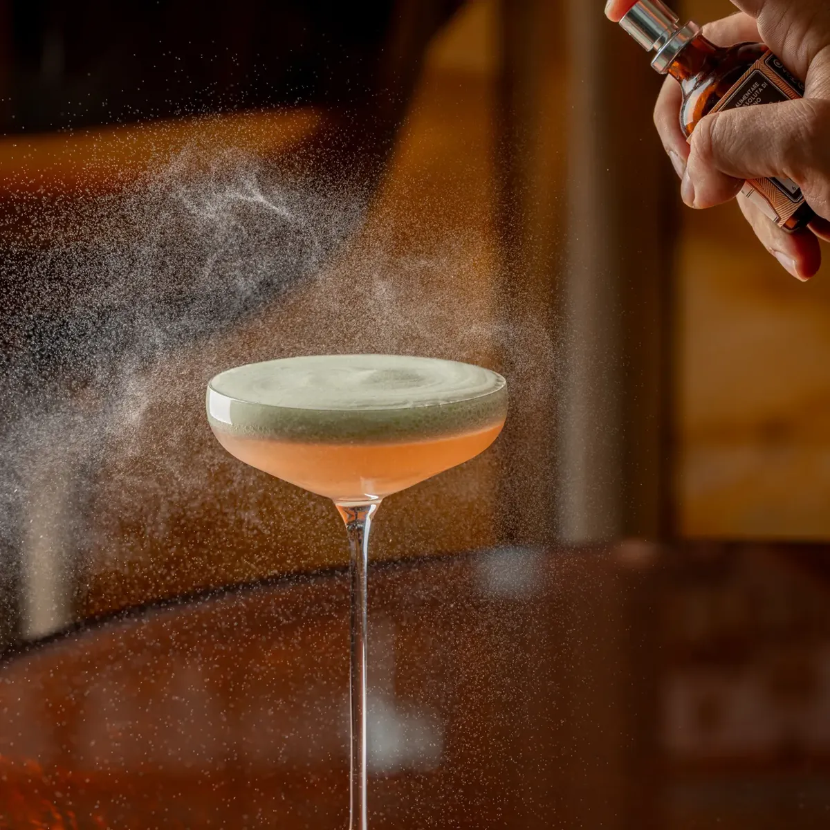 A hand sprays a fine mist over a pale pink cocktail in a coupe glass on a polished wooden bar.