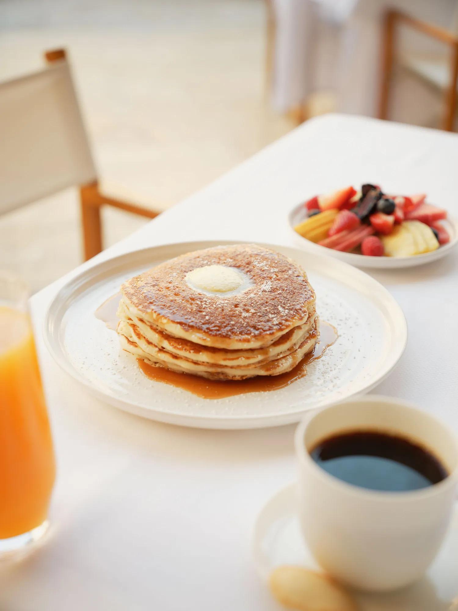 Table with white linen, fresh fruits, coffee, orange juice, and fluffy pancakes.