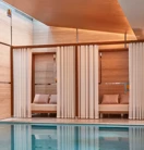 Indoor pool with soft lighting, featuring private cabanas framed by white curtains and furnished with cushioned seating against warm-toned wooden walls.