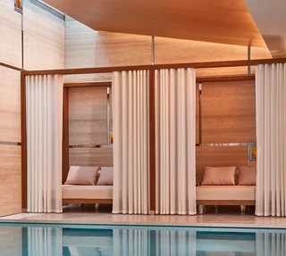 Indoor pool with soft lighting, featuring private cabanas framed by white curtains and furnished with cushioned seating against warm-toned wooden walls.