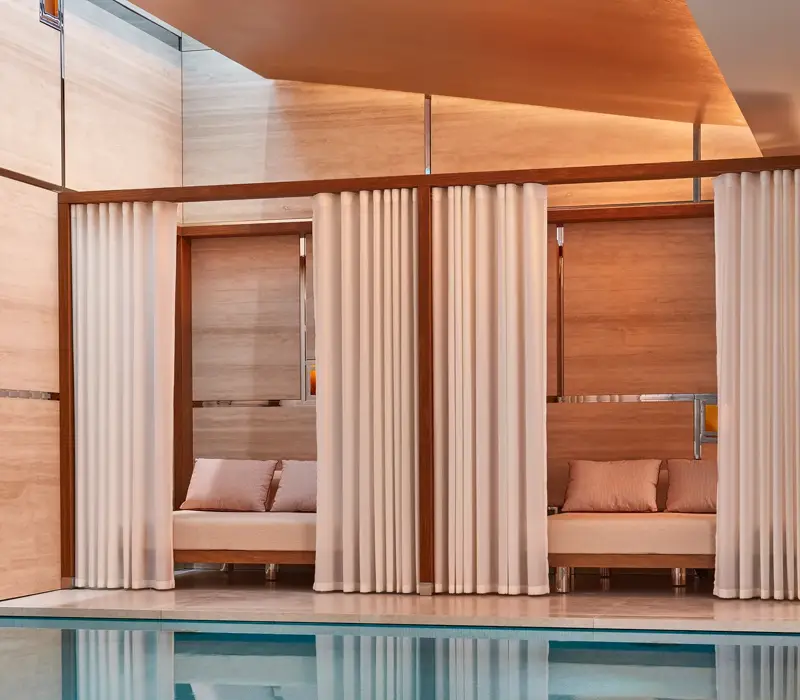Indoor pool with soft lighting, featuring private cabanas framed by white curtains and furnished with cushioned seating against warm-toned wooden walls.