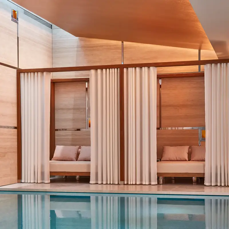 Indoor pool with soft lighting, featuring private cabanas framed by white curtains and furnished with cushioned seating against warm-toned wooden walls.