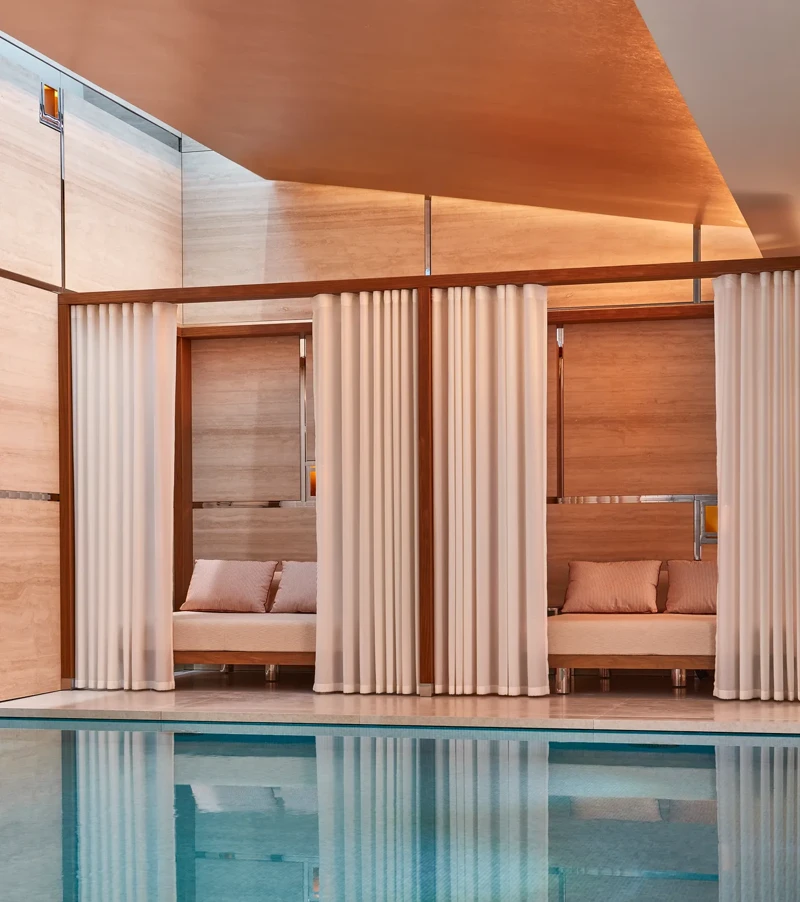 Indoor pool with soft lighting, featuring private cabanas framed by white curtains and furnished with cushioned seating against warm-toned wooden walls.