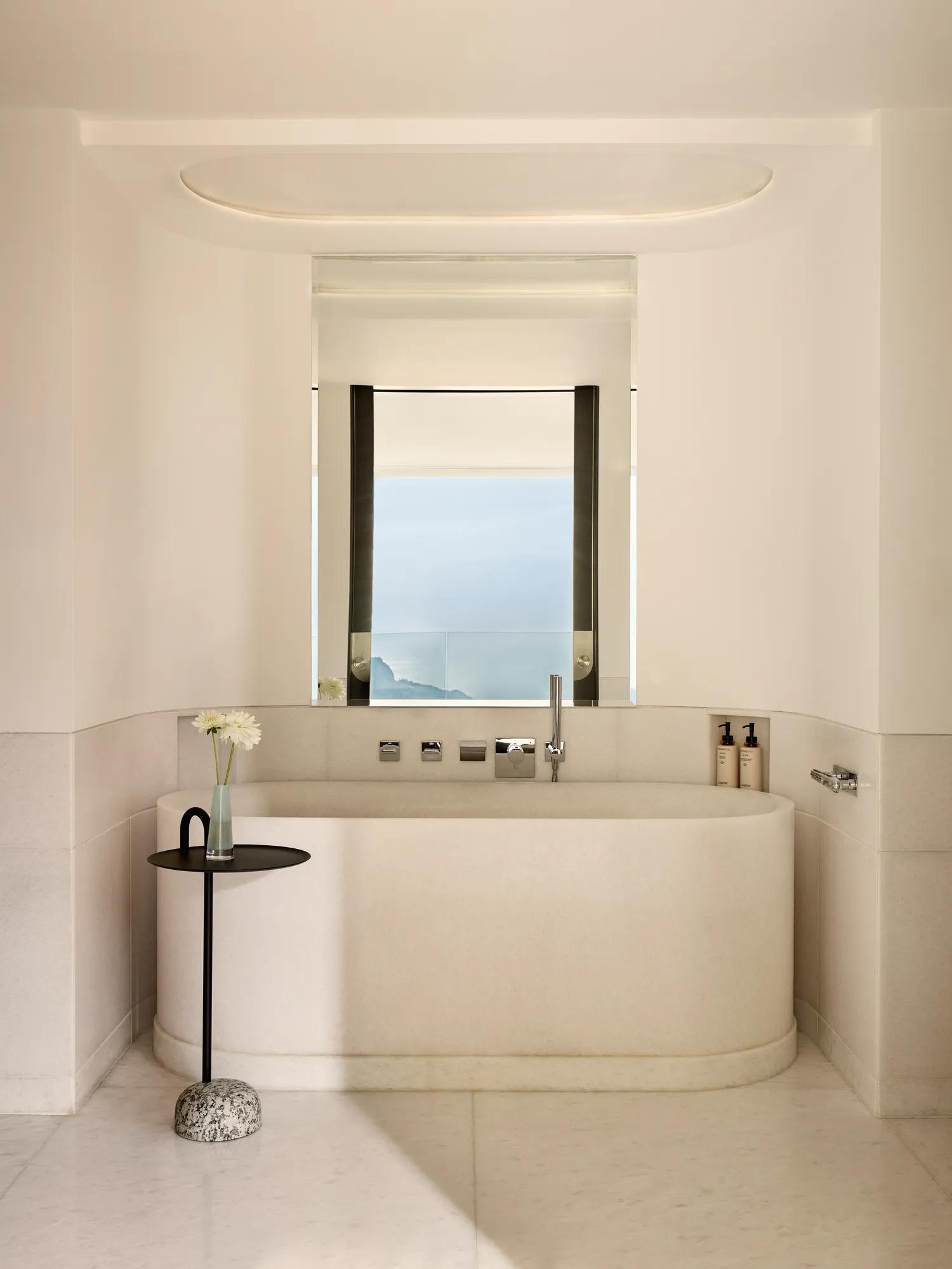 Minimalist bathroom with freestanding bathtub beneath a window, offering a serene sea view, complemented by a small side table with a single flower.