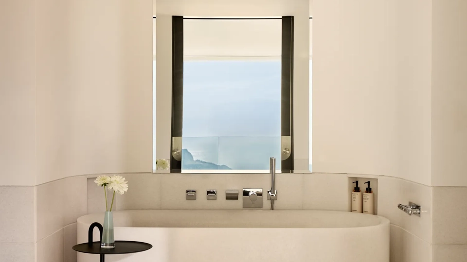 Minimalist bathroom with freestanding bathtub beneath a window, offering a serene sea view, complemented by a small side table with a single flower.