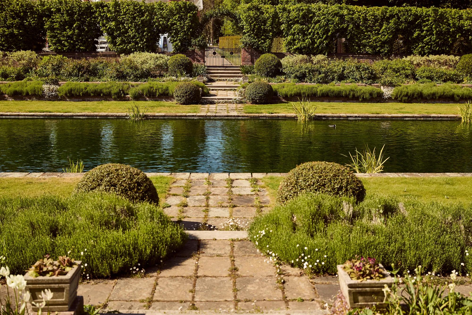 Formal garden with stone pathway leading to a rectangular pond, surrounded by manicured shrubs, flower beds, and lush greenery.