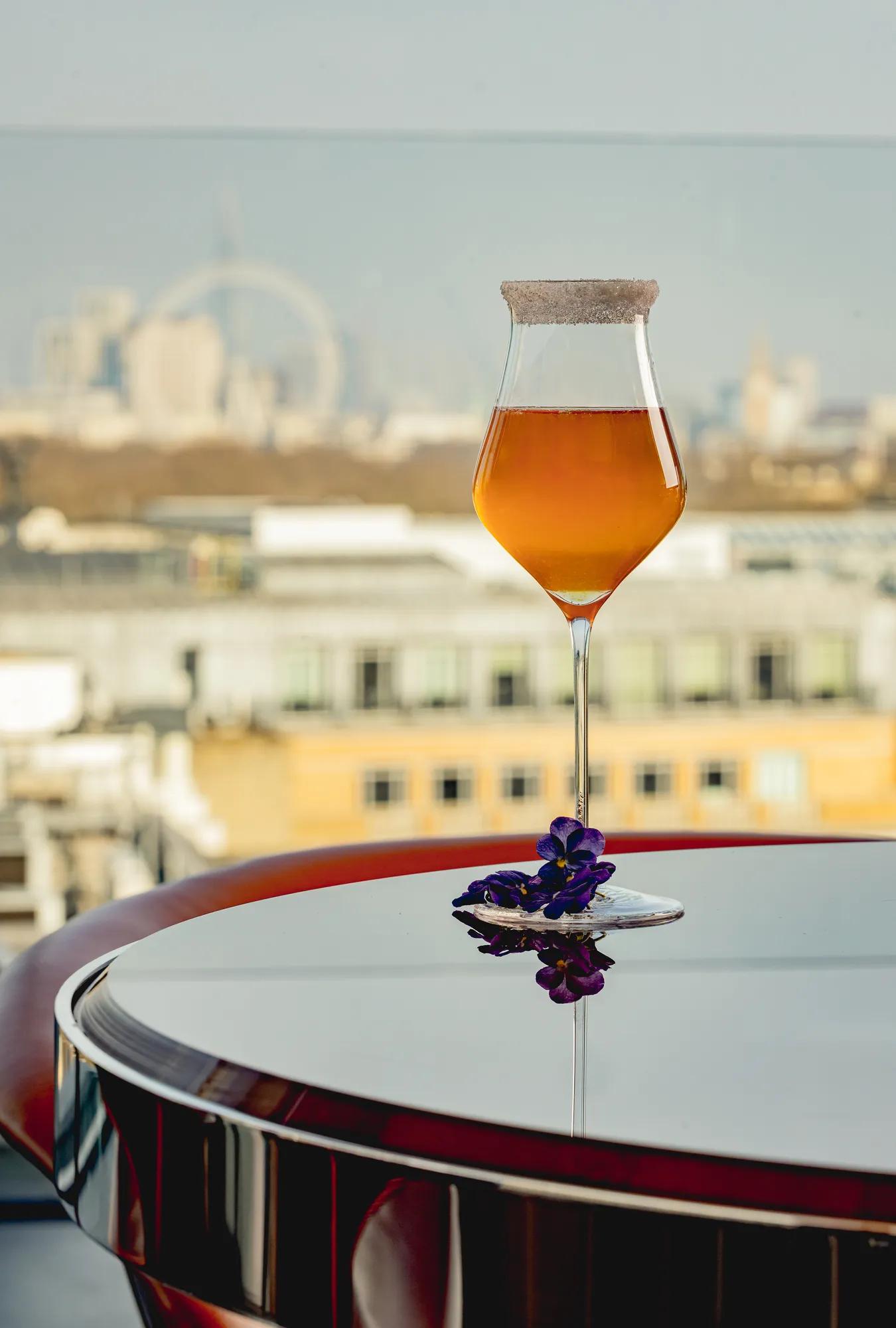 An amber cocktail with a sugared rim on a reflective table, garnished with purple flowers and a city skyline view.