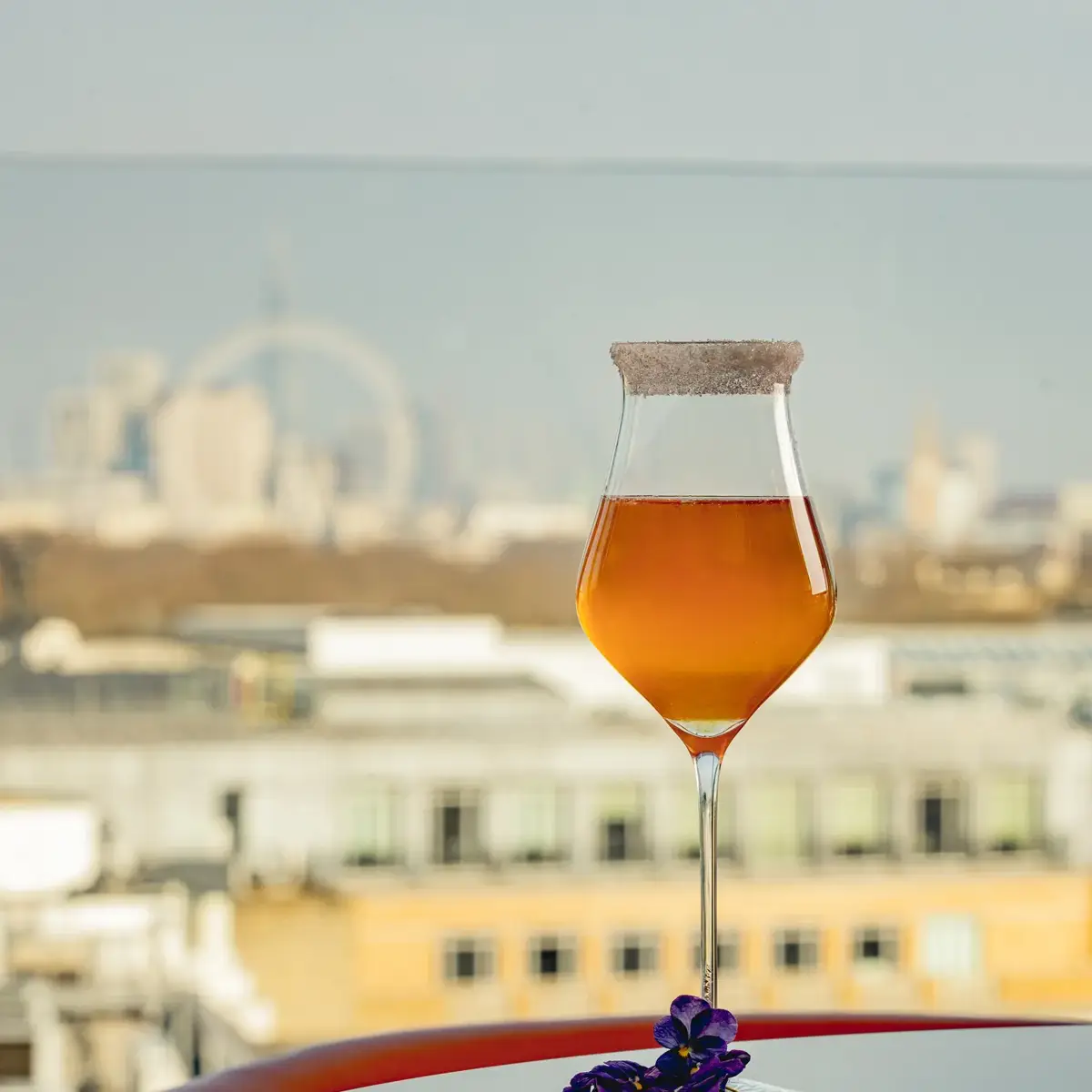 An amber cocktail with a sugared rim on a reflective table, garnished with purple flowers and a city skyline view.