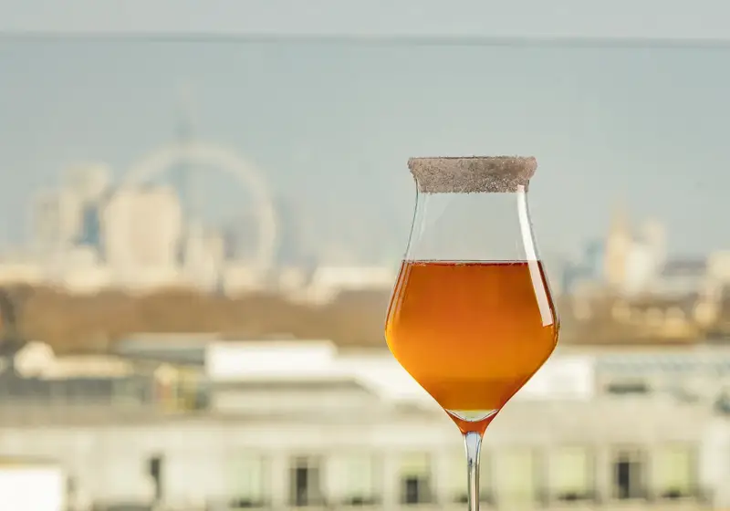 An amber cocktail with a sugared rim on a reflective table, garnished with purple flowers and a city skyline view.