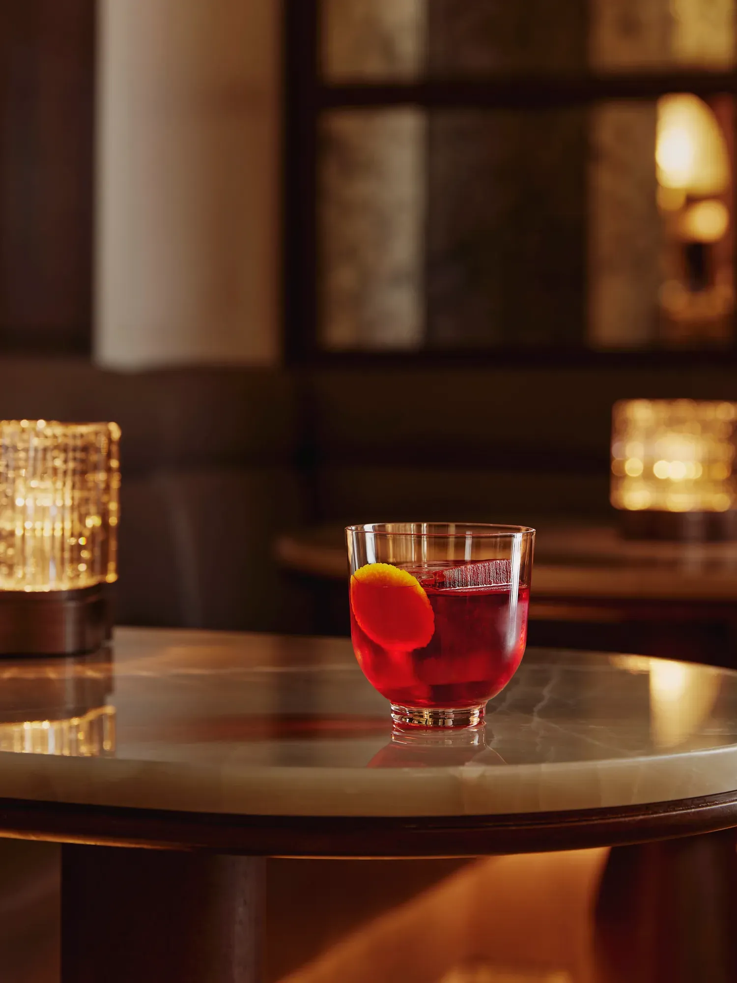 Short red cocktail with orange peel garnish on a marble table; warm candlelight bokeh behind.