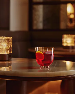 Short red cocktail with orange peel garnish on a marble table; warm candlelight bokeh behind.
