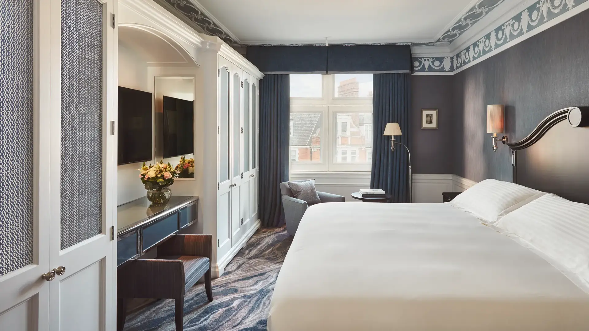 Bedroom with large bed, blue walls and drapes, white wardrobe cabinets, and a seating area by the window.