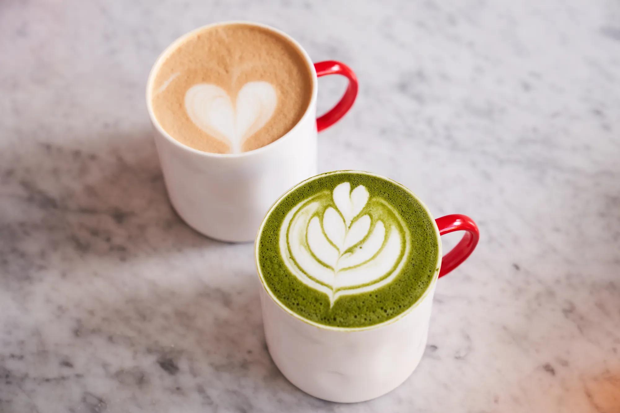 Two lattes in white mugs with red handles, one matcha and one coffee, topped with heart latte art.