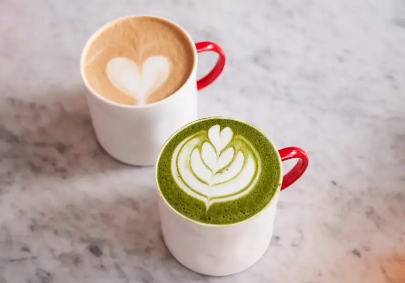Two lattes in white mugs with red handles, one matcha and one coffee, topped with heart latte art.