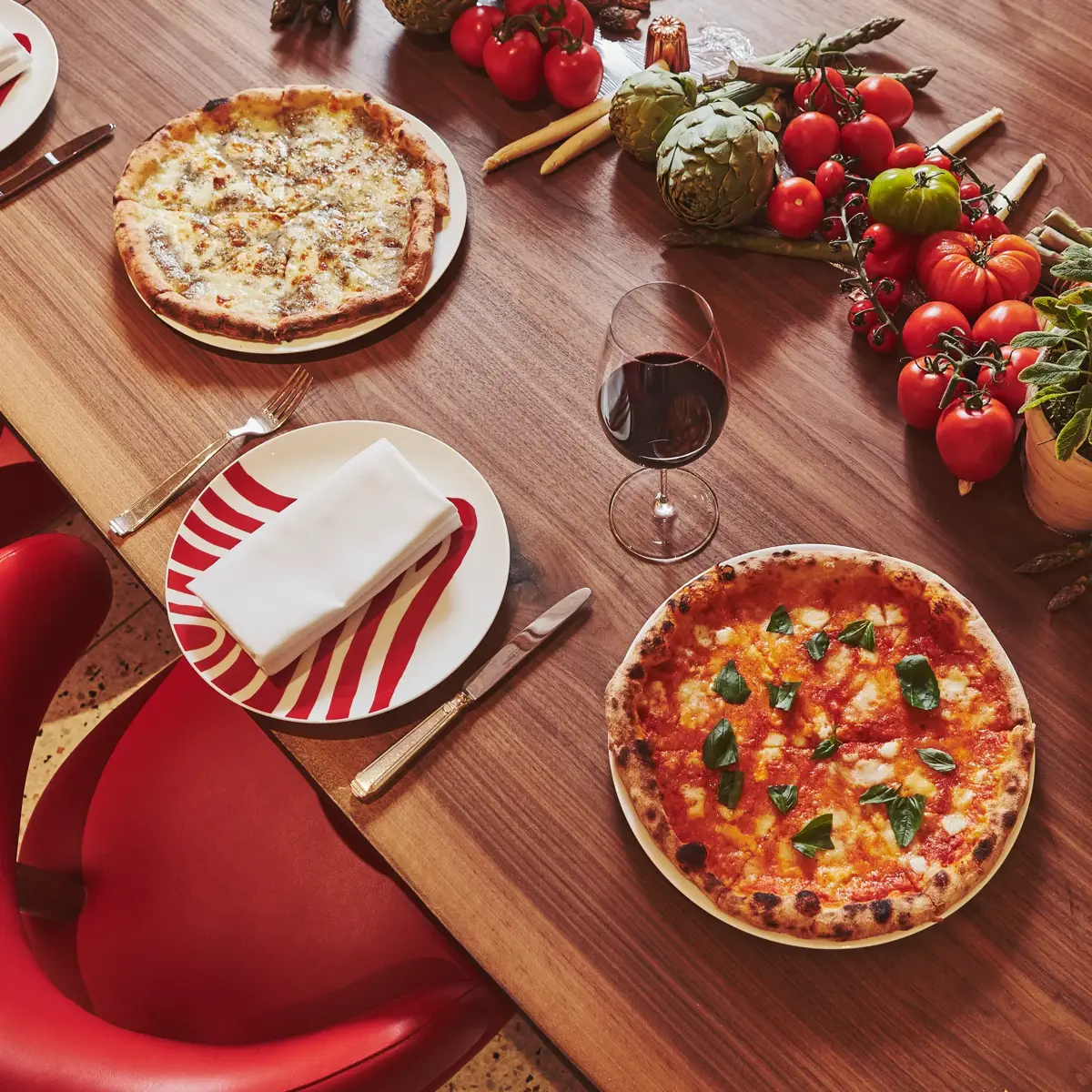 Two pizzas, a glass of red wine, and fresh tomatoes and vegetables displayed on a wooden table set with red chairs and striped plates.
