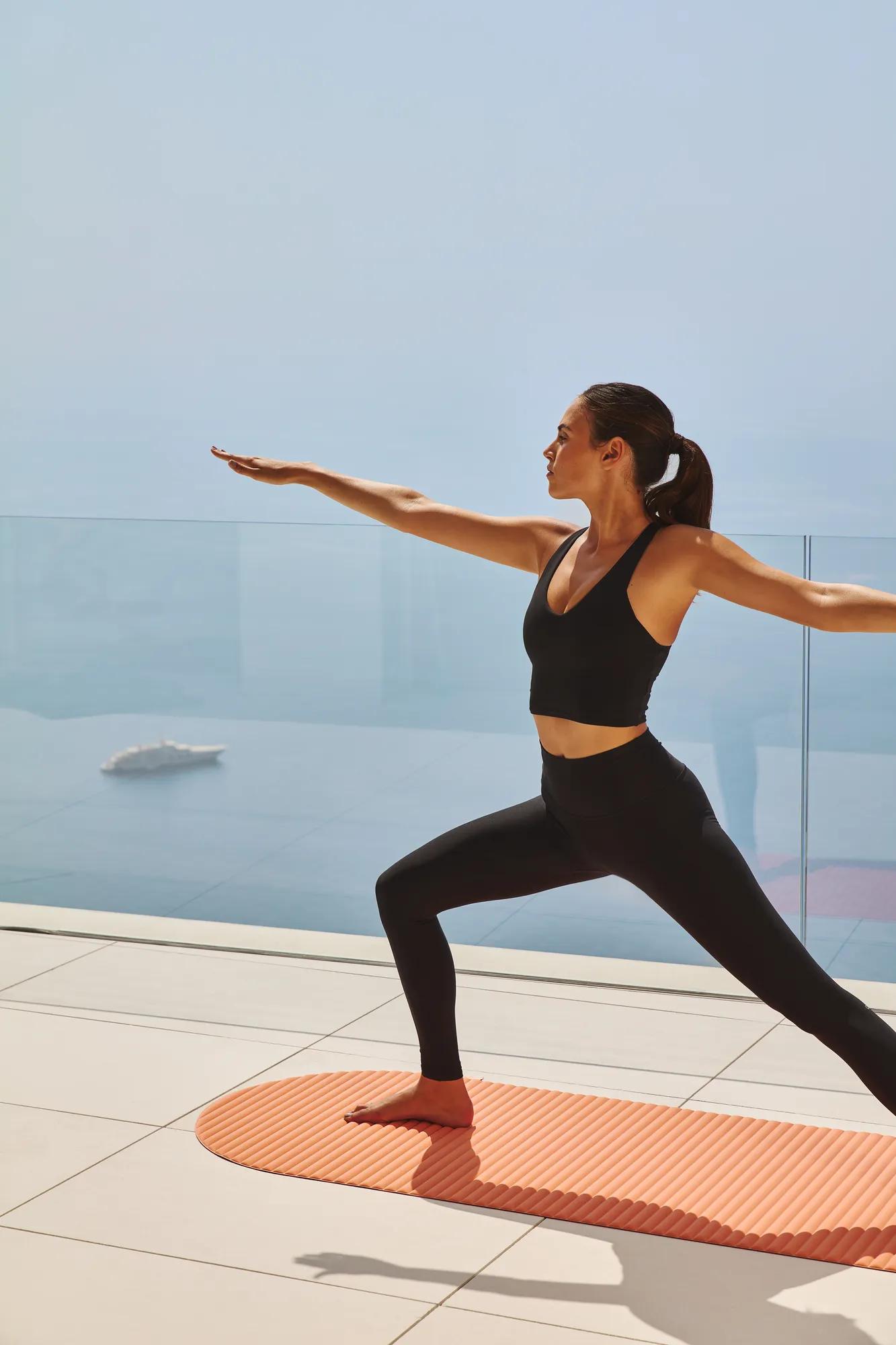 Person practicing yoga in a warrior pose on an orange mat, with the backdrop of a clear, open view overlooking the sea and a modern glass railing.