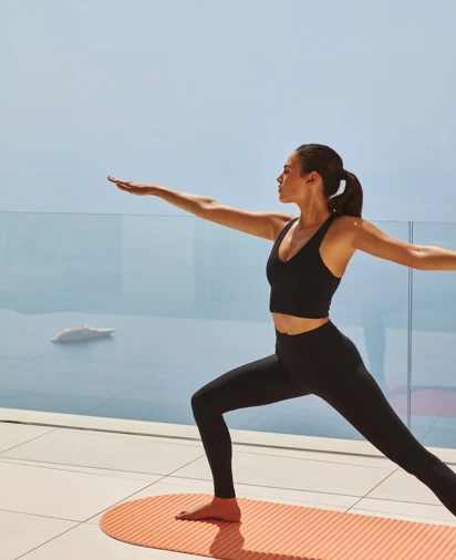 Person practicing yoga in a warrior pose on an orange mat, with the backdrop of a clear, open view overlooking the sea and a modern glass railing.