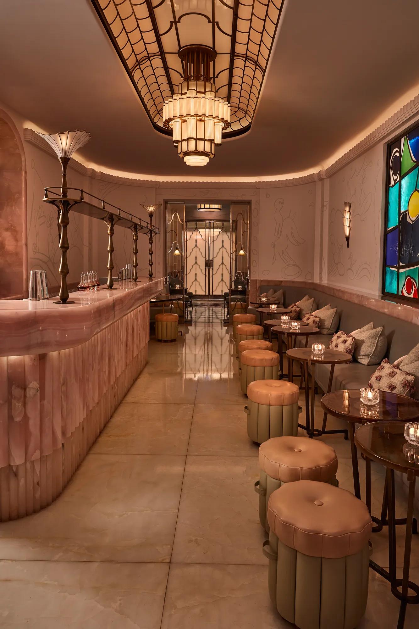 Art Deco bar interior with marble counter, upholstered stools and small tables, featuring stained glass window, decorative lighting and soft ambient glow.