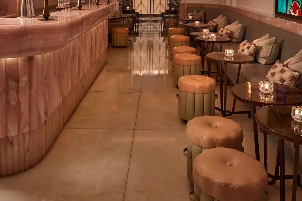 Art Deco bar interior with marble counter, upholstered stools and small tables, featuring stained glass window, decorative lighting and soft ambient glow.