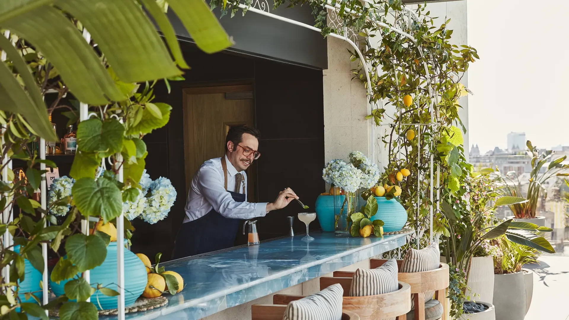 Rooftop bar with blue counter, wooden stools, and lush greenery, bartender preparing cocktails under sunshine.