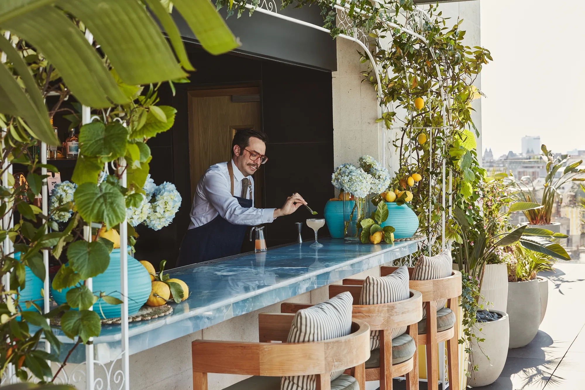 Rooftop bar with blue counter, wooden stools, and lush greenery, bartender preparing cocktails under sunshine.
