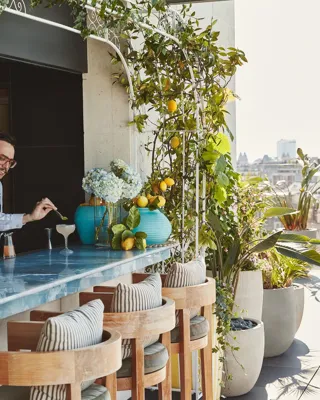 Rooftop bar with blue counter, wooden stools, and lush greenery, bartender preparing cocktails under sunshine.