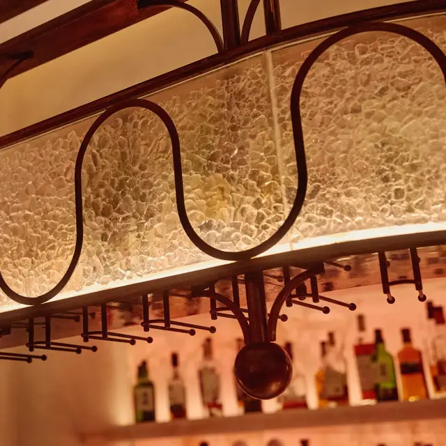 Close-up of a textured glass and bronze bar light fixture with warm backlighting and bottles displayed below.