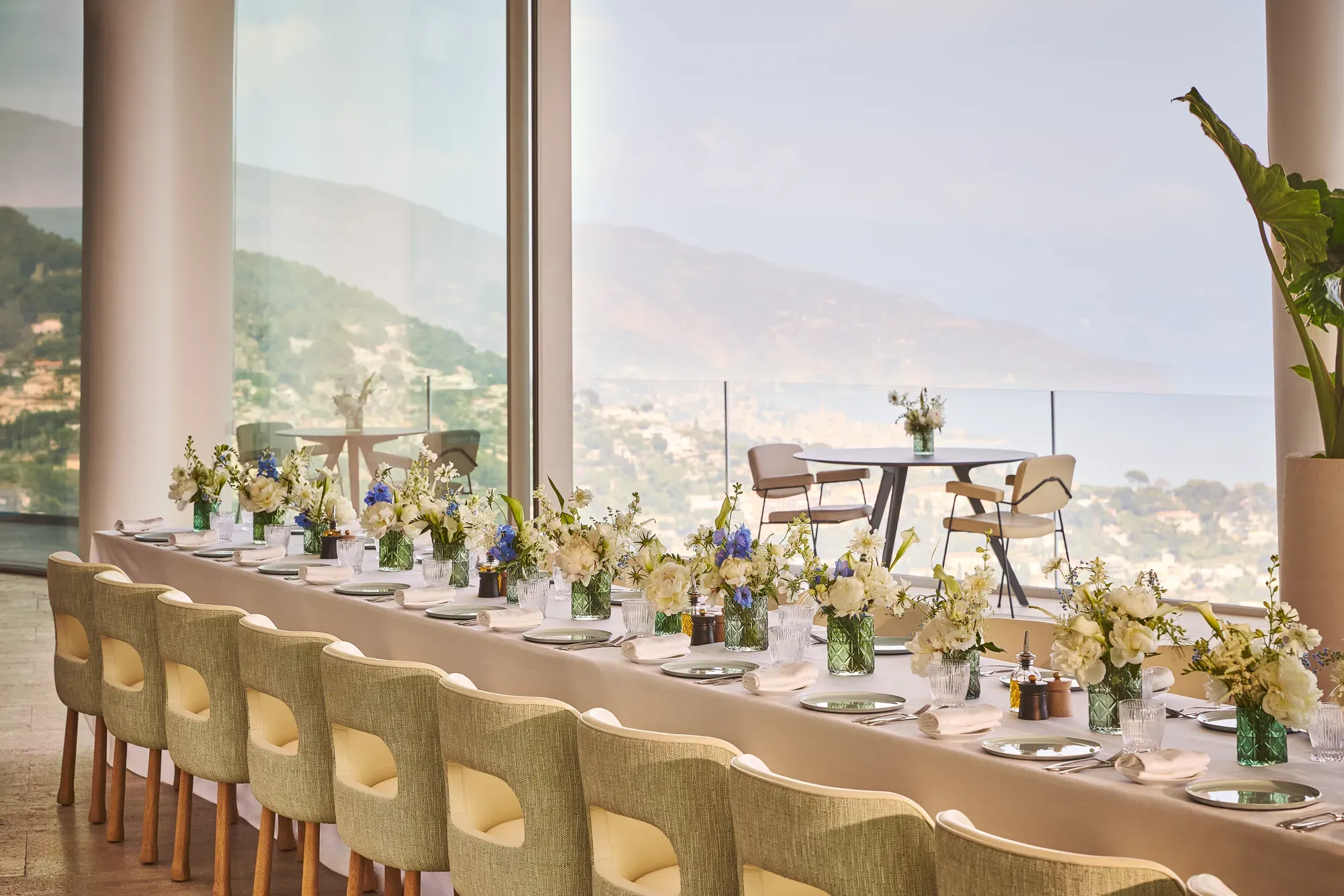 Longue table dressée avec nappes neutres, compositions florales, chaises rembourrées et vue panoramique mer et montagnes.