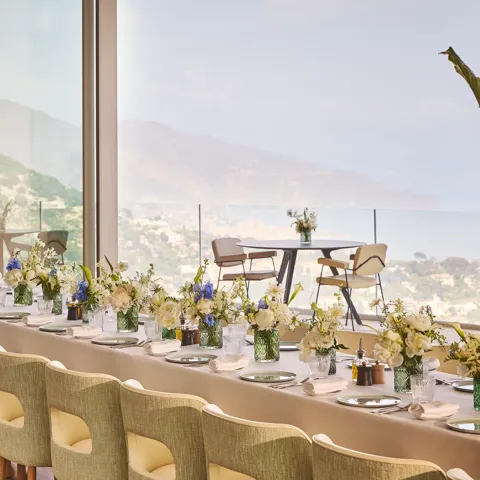 Longue table dressée avec nappes neutres, compositions florales, chaises rembourrées et vue panoramique mer et montagnes.