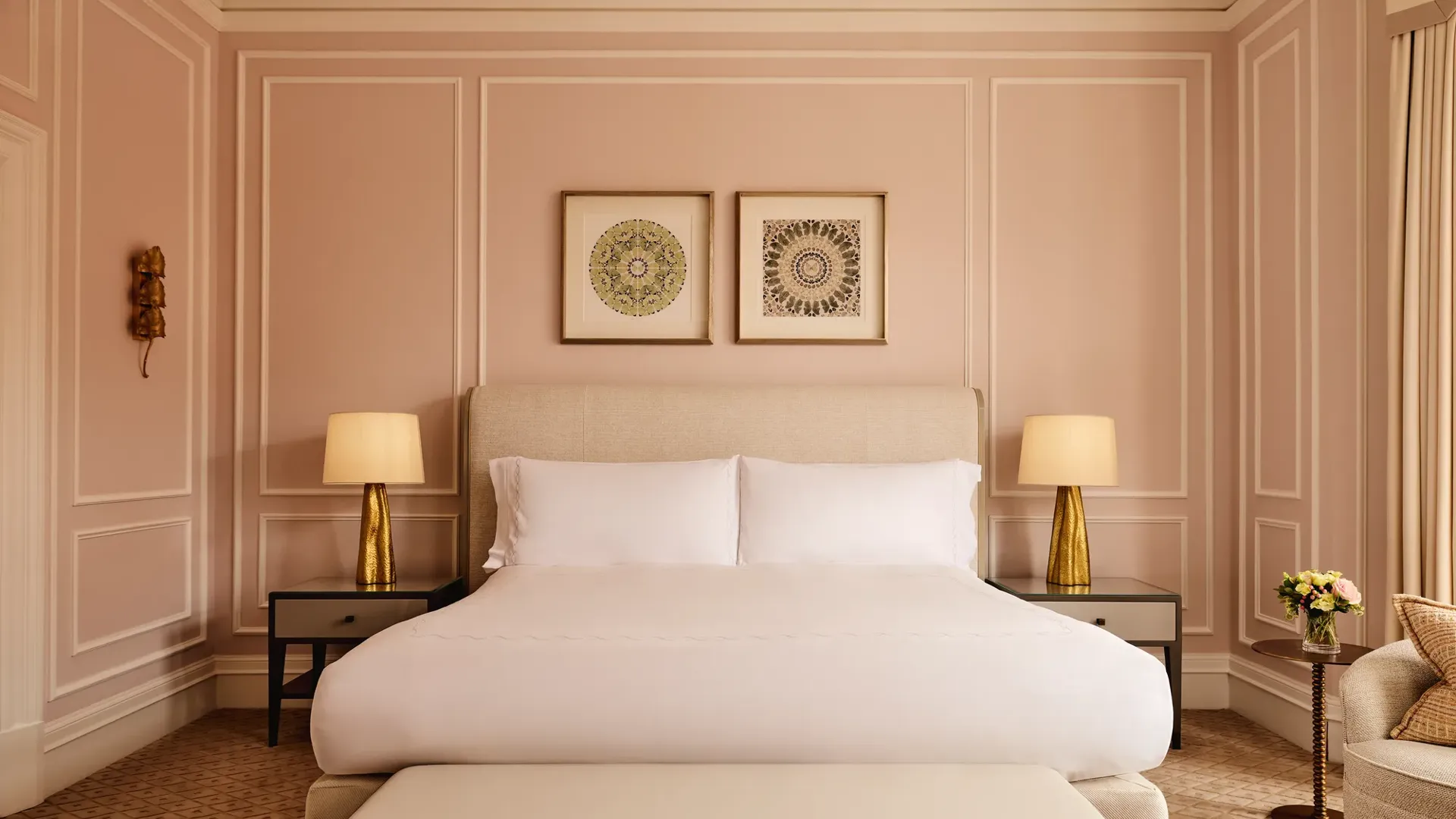 Elegant bedroom with blush-pink panelled walls, a king-size bed with crisp white linens, two golden lamps on bedside tables, and framed artwork above the headboard.