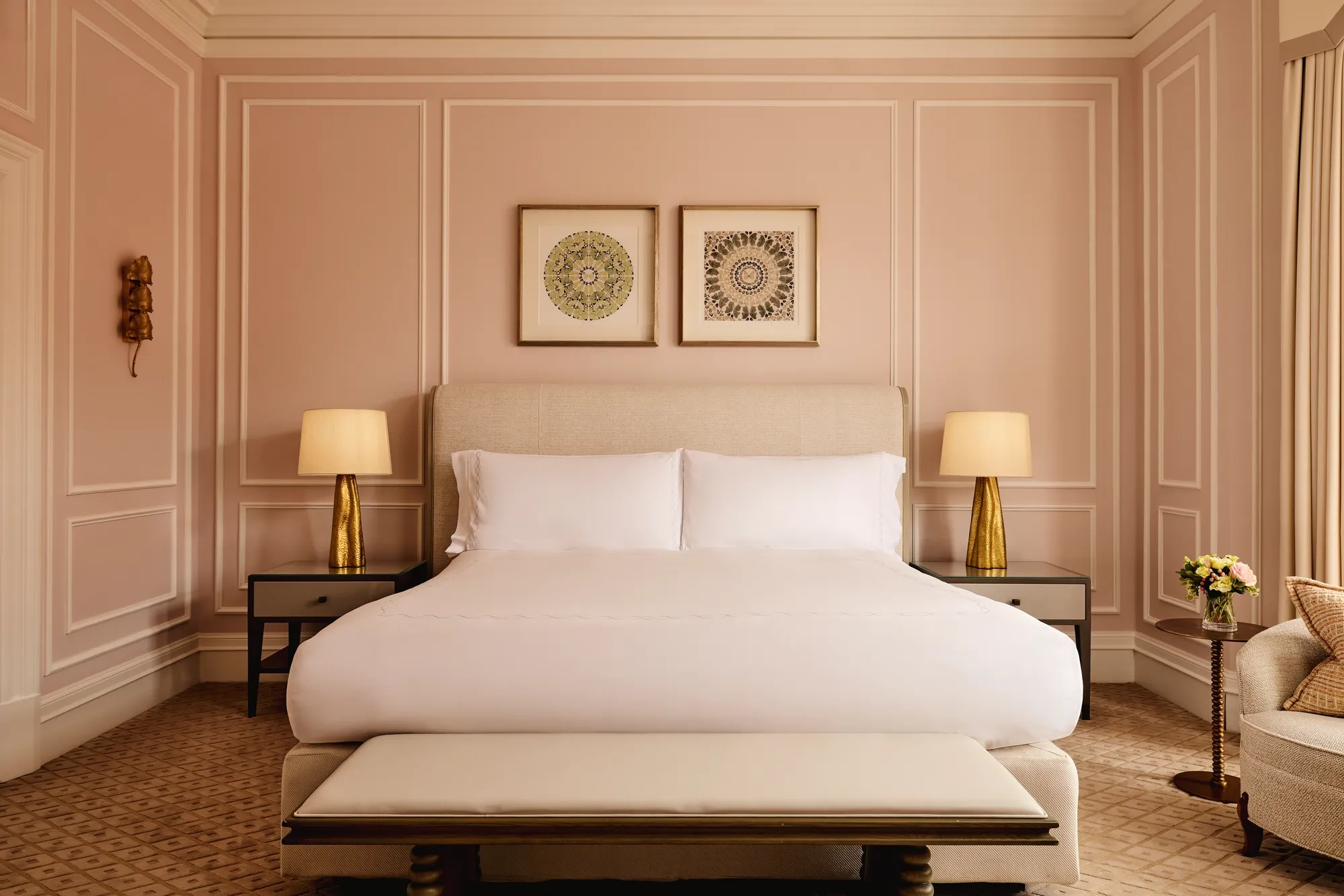 Elegant bedroom with blush-pink panelled walls, a king-size bed with crisp white linens, two golden lamps on bedside tables, and framed artwork above the headboard.